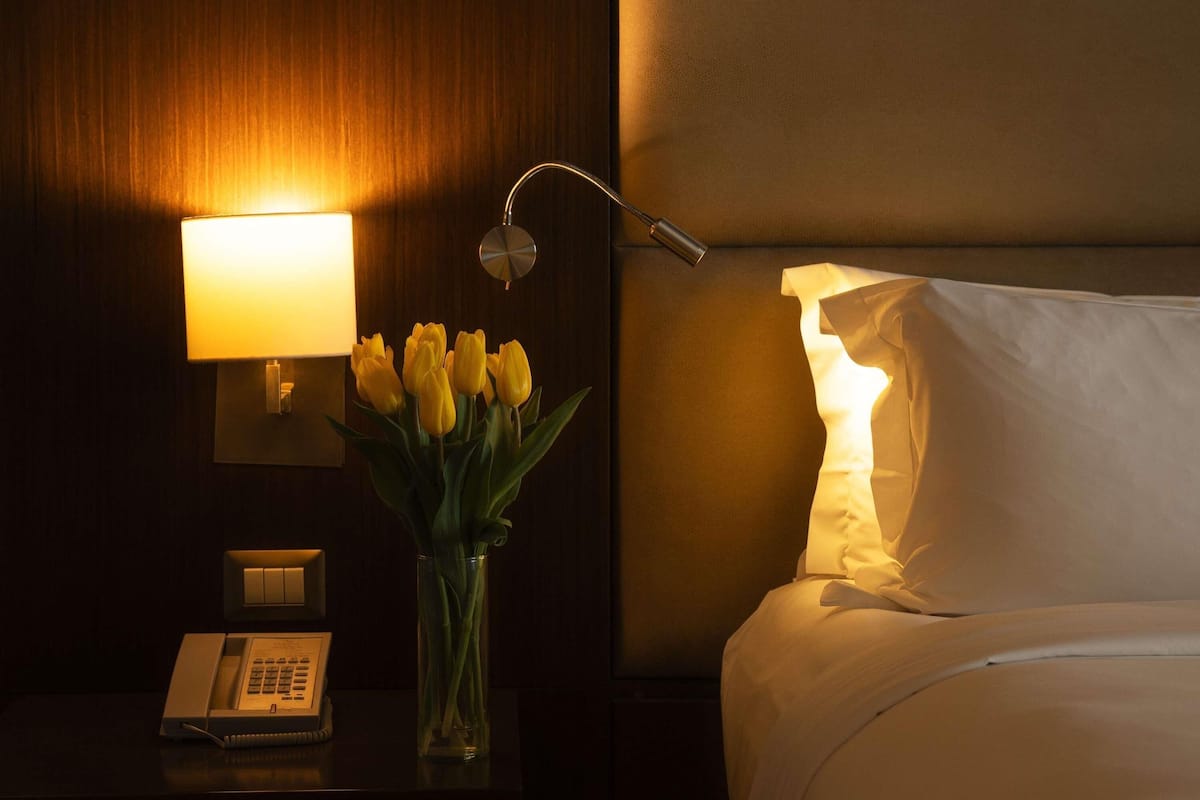 Hotellihuone, jossa on yöpöydällä lamppu, puhelin, maljakko kukkineen ja sängyssä tyyny.