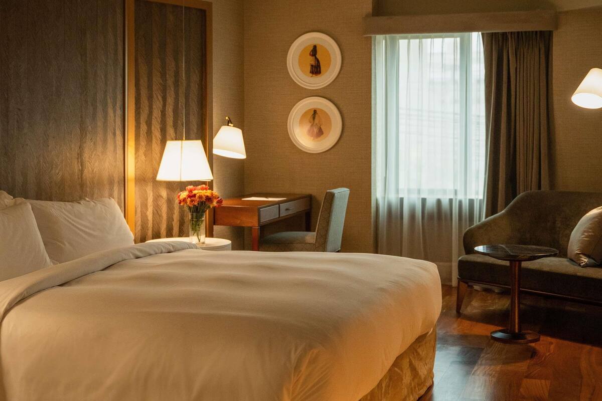 A hotel room with a large bed, a desk with a lamp, a chair, and a view of the outdoors through a window.