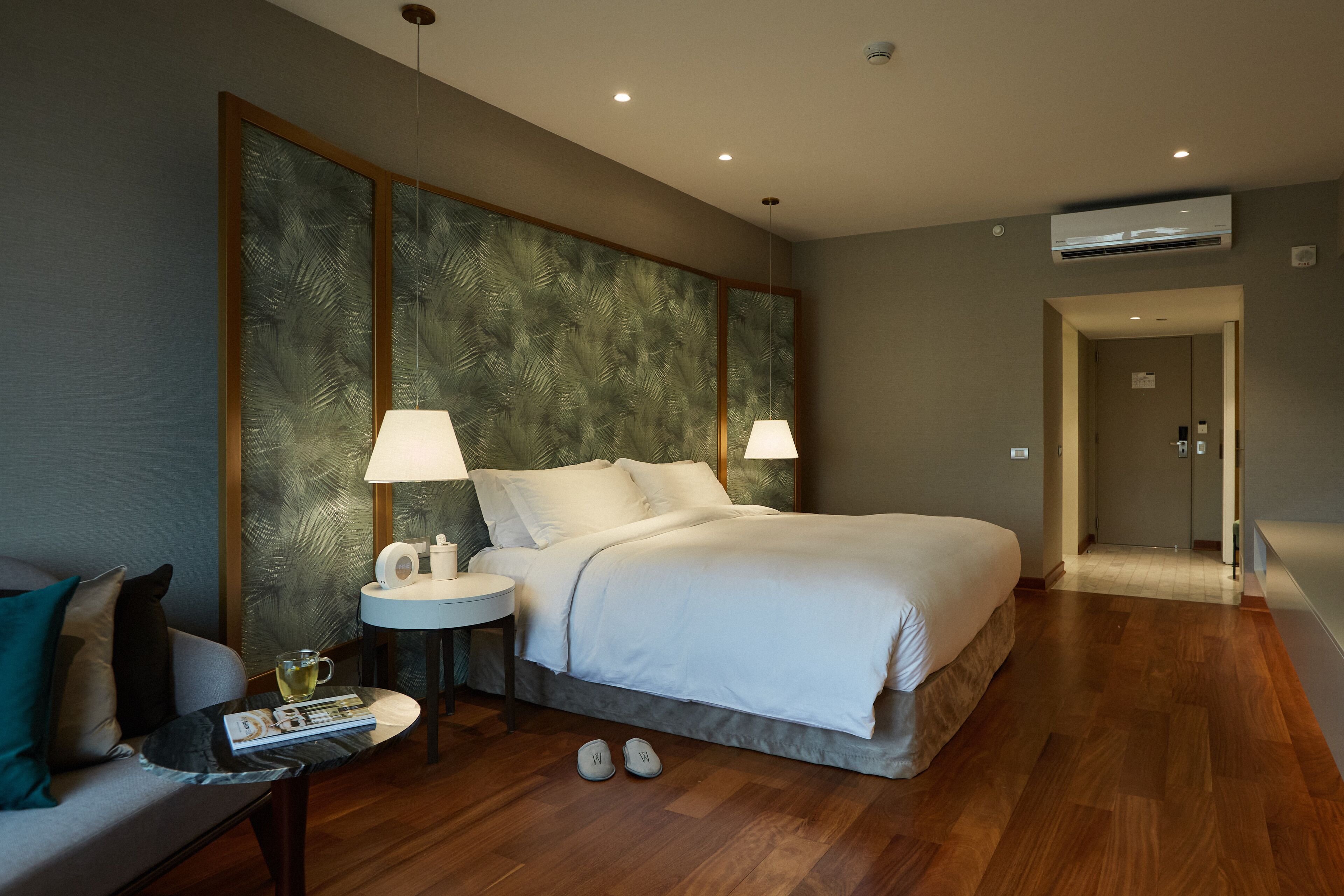 Een hotelkamer met een groot bed, twee bedlampen, een kleine tafel met een lamp, een bank en een paar slippers op de vloer.