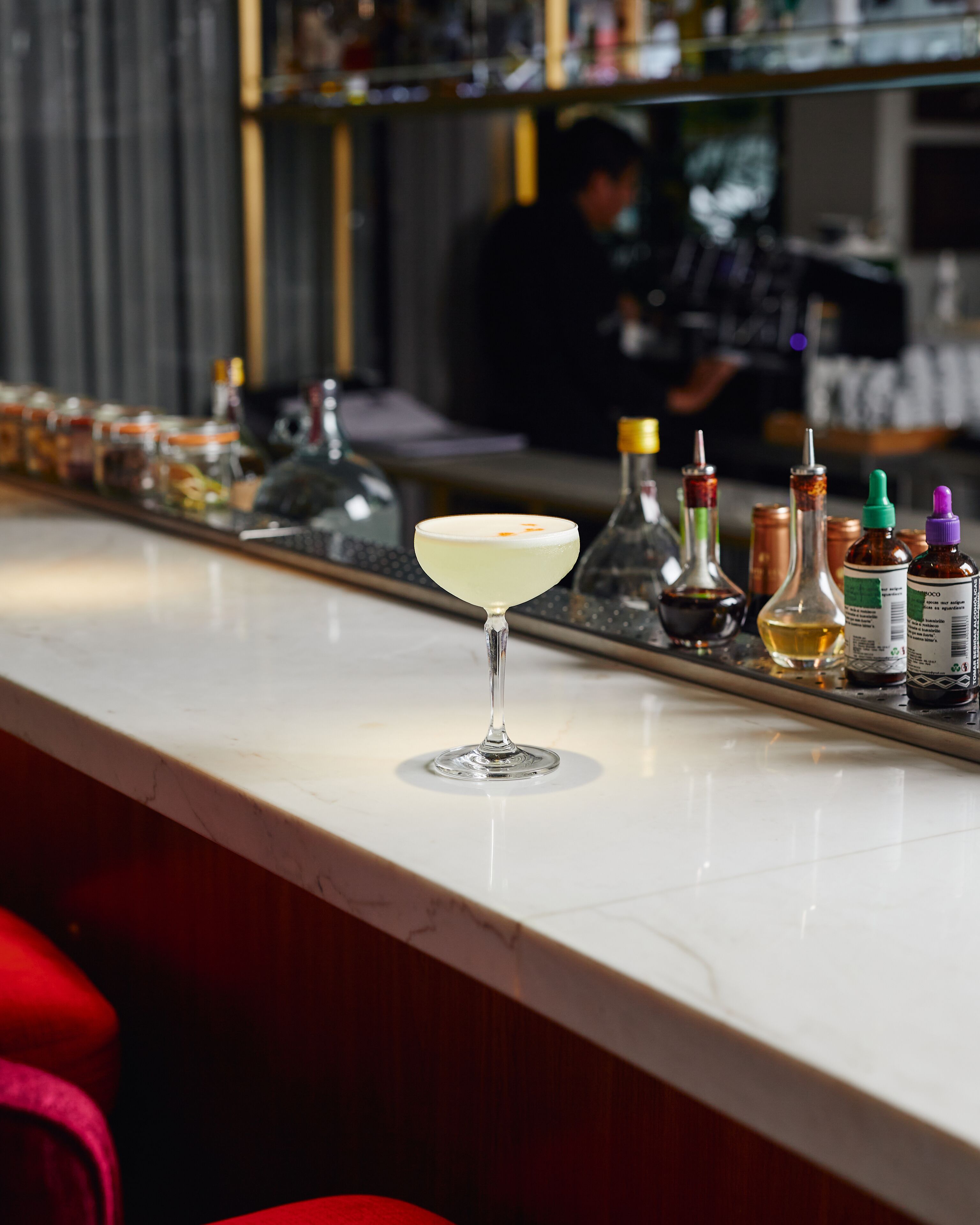 A bar counter with a martini glass in the foreground, various bottles and condiments on the counter, and a blurred figure in the background.