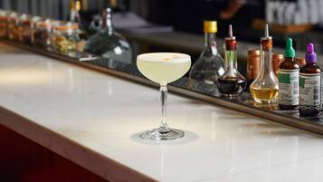 A bar counter with a martini glass in the foreground, various bottles and condiments on the counter, and a blurred figure in the background.