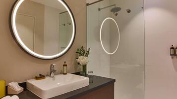 Un baño con un espejo redondo, un lavabo rectangular, una cabina de ducha de cristal y un florero con flores en la encimera.