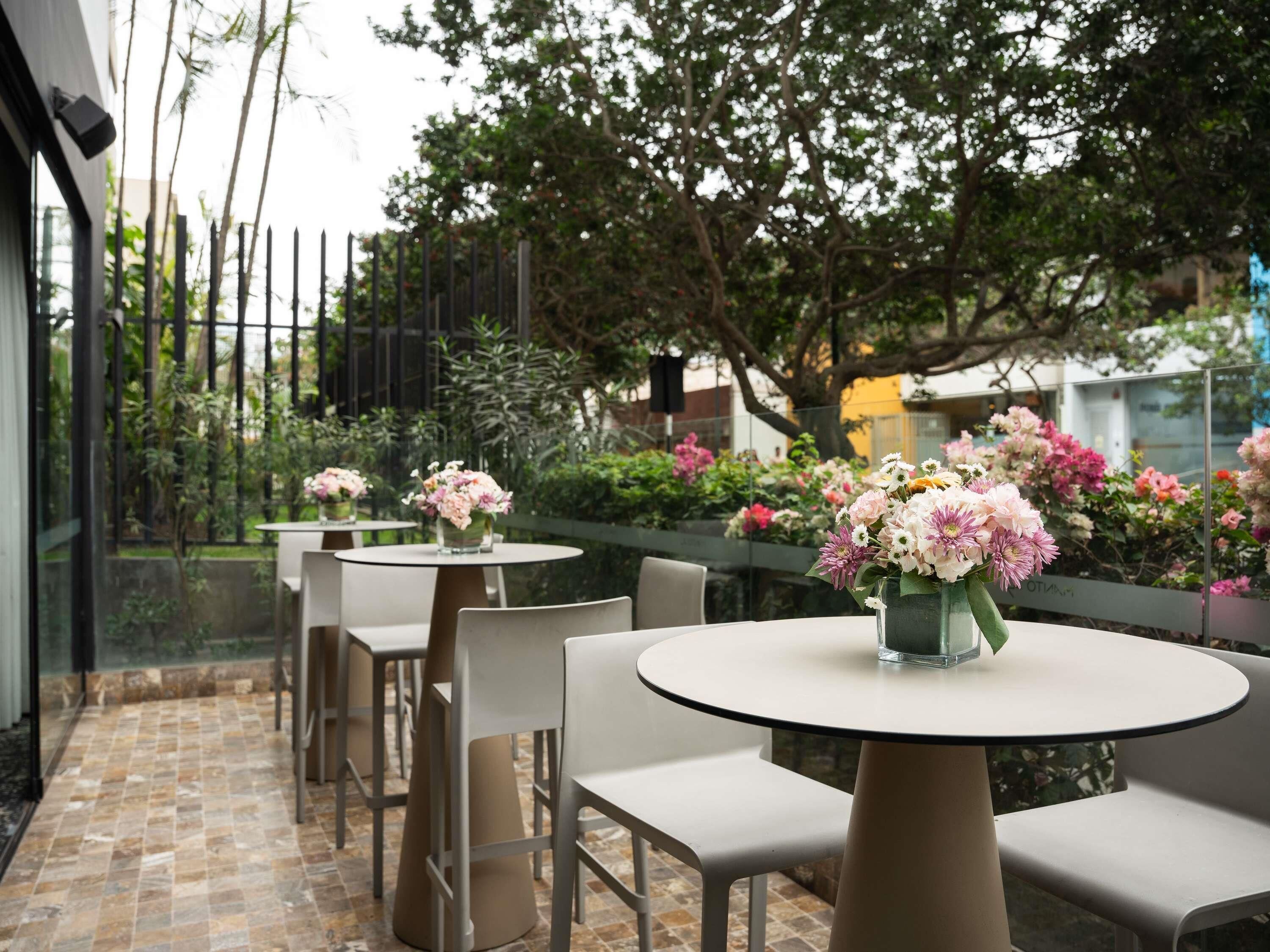 A patio area with round tables and chairs, surrounded by a glass railing, with a view of a garden and trees.