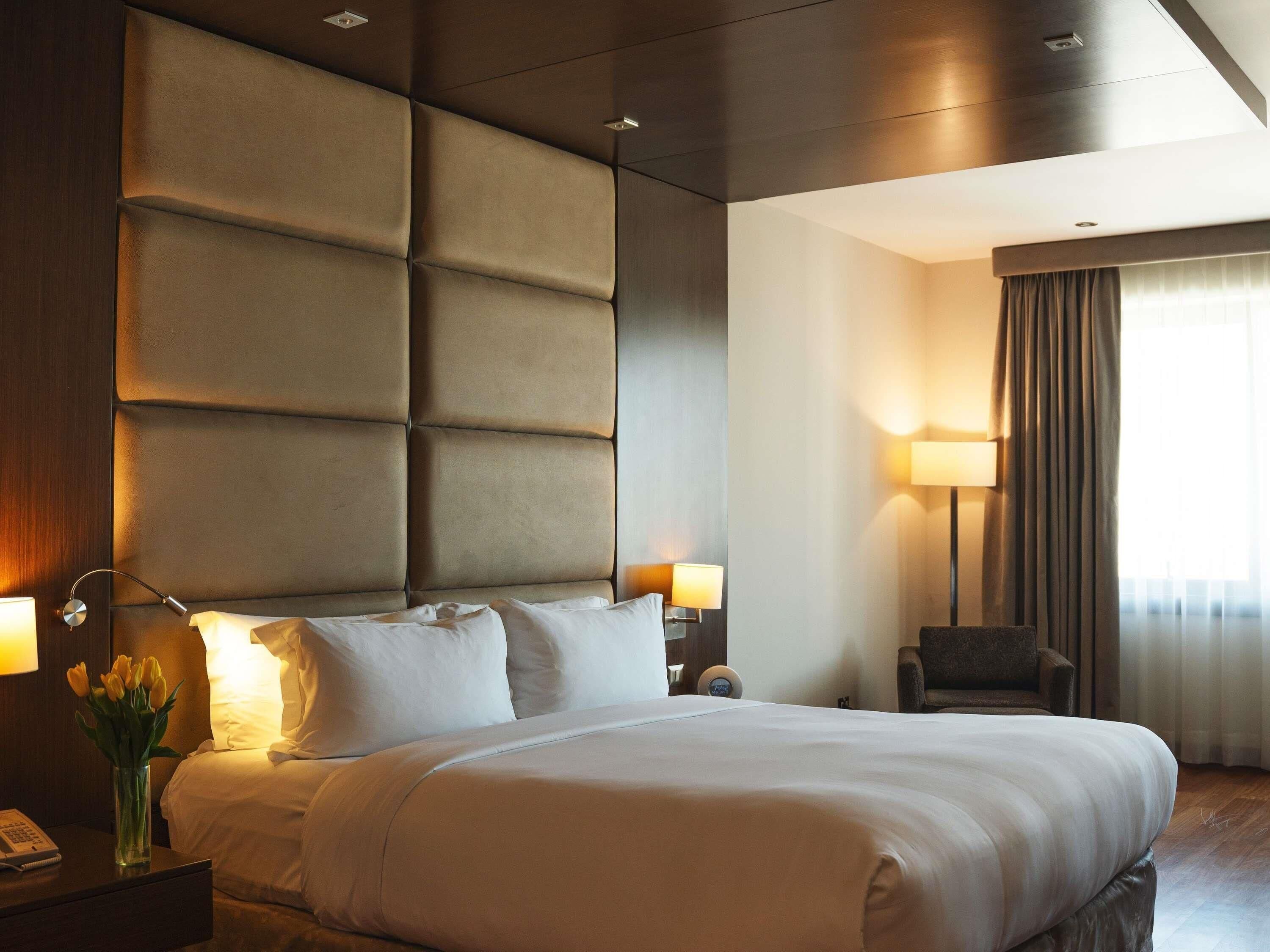 Habitación de hotel con una cama grande, una mesita de noche con lámpara, una silla y una ventana con cortinas.