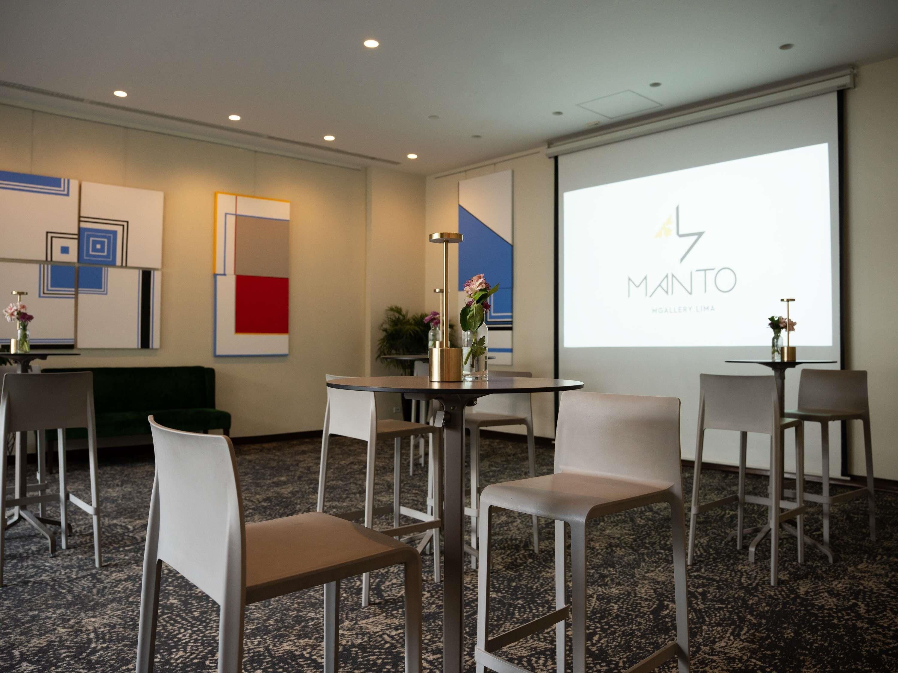 Un salón de conferencias con una pantalla de proyector mostrando 'MANTO' y 'MOULERY LIMA', sillas blancas, una mesa redonda y arte abstracto en la pared.