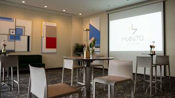 Un salón de conferencias con una pantalla de proyector mostrando 'MANTO' y 'MOULERY LIMA', sillas blancas, una mesa redonda y arte abstracto en la pared.