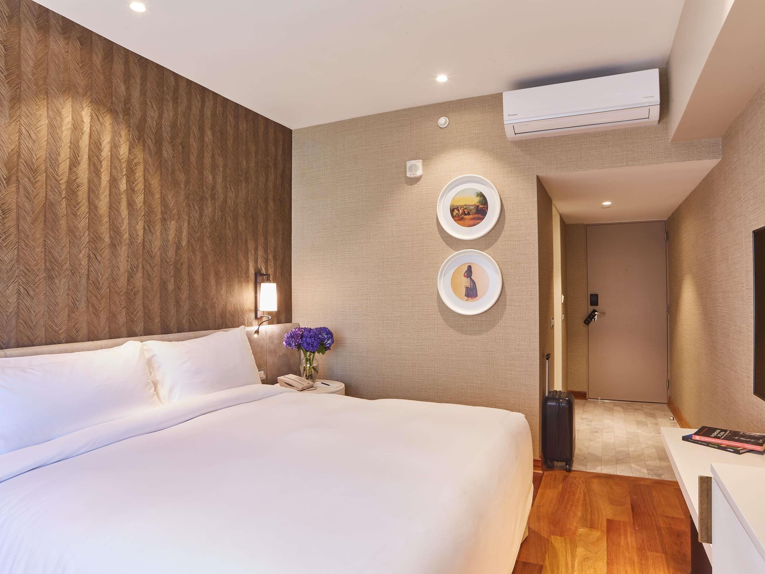 Una habitación de hotel con una cama grande, aire acondicionado montado en la pared, una mesita de noche con un jarrón de flores y una puerta que da a un baño.