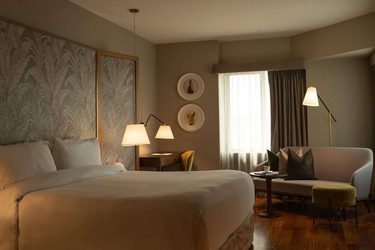 A hotel room with a large bed, two bedside lamps, a desk, a chair, and a window with curtains.