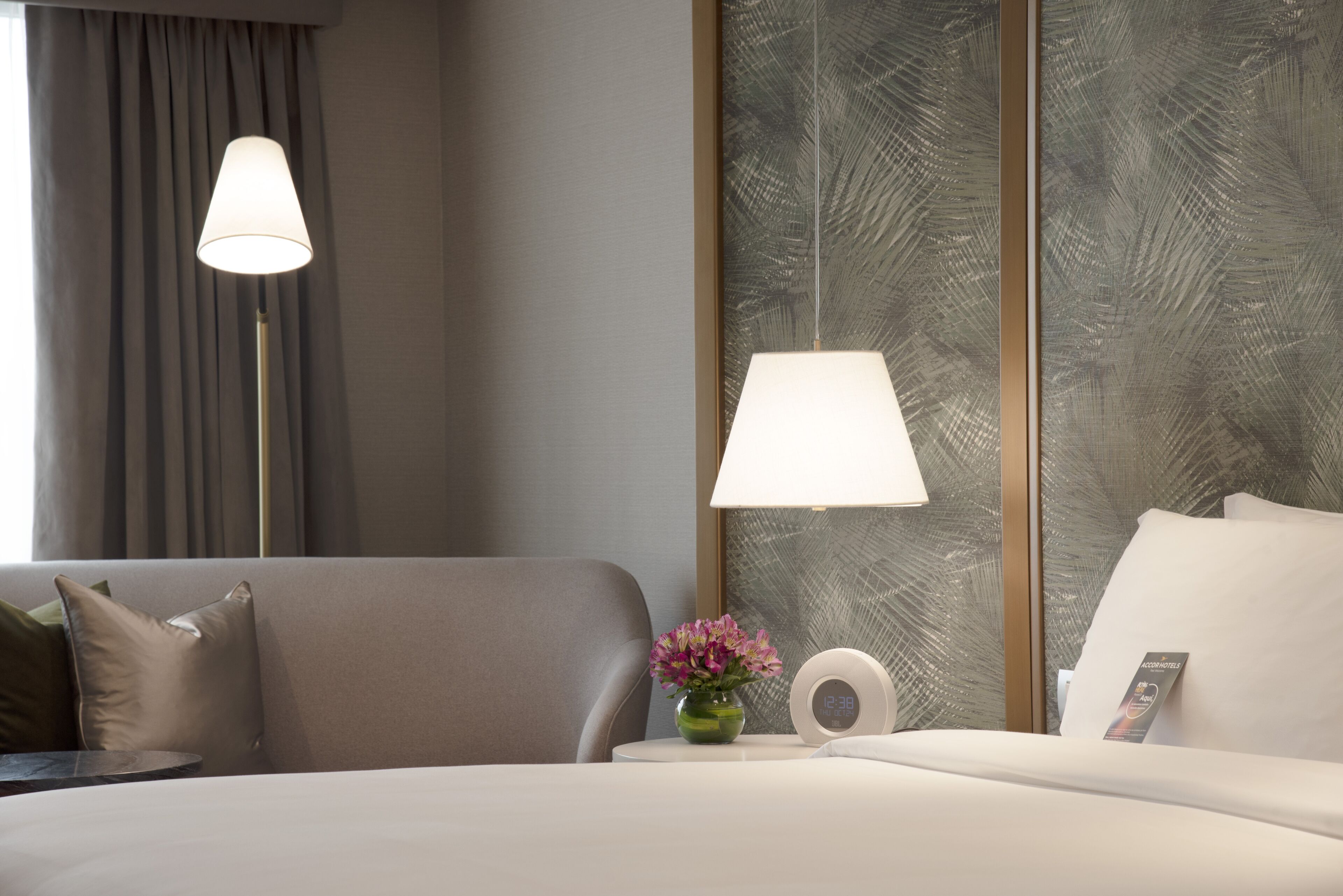 Una habitación de hotel moderna con una cama, una mesita de noche con un reloj y un jarrón de flores, y dos lámparas colgantes.