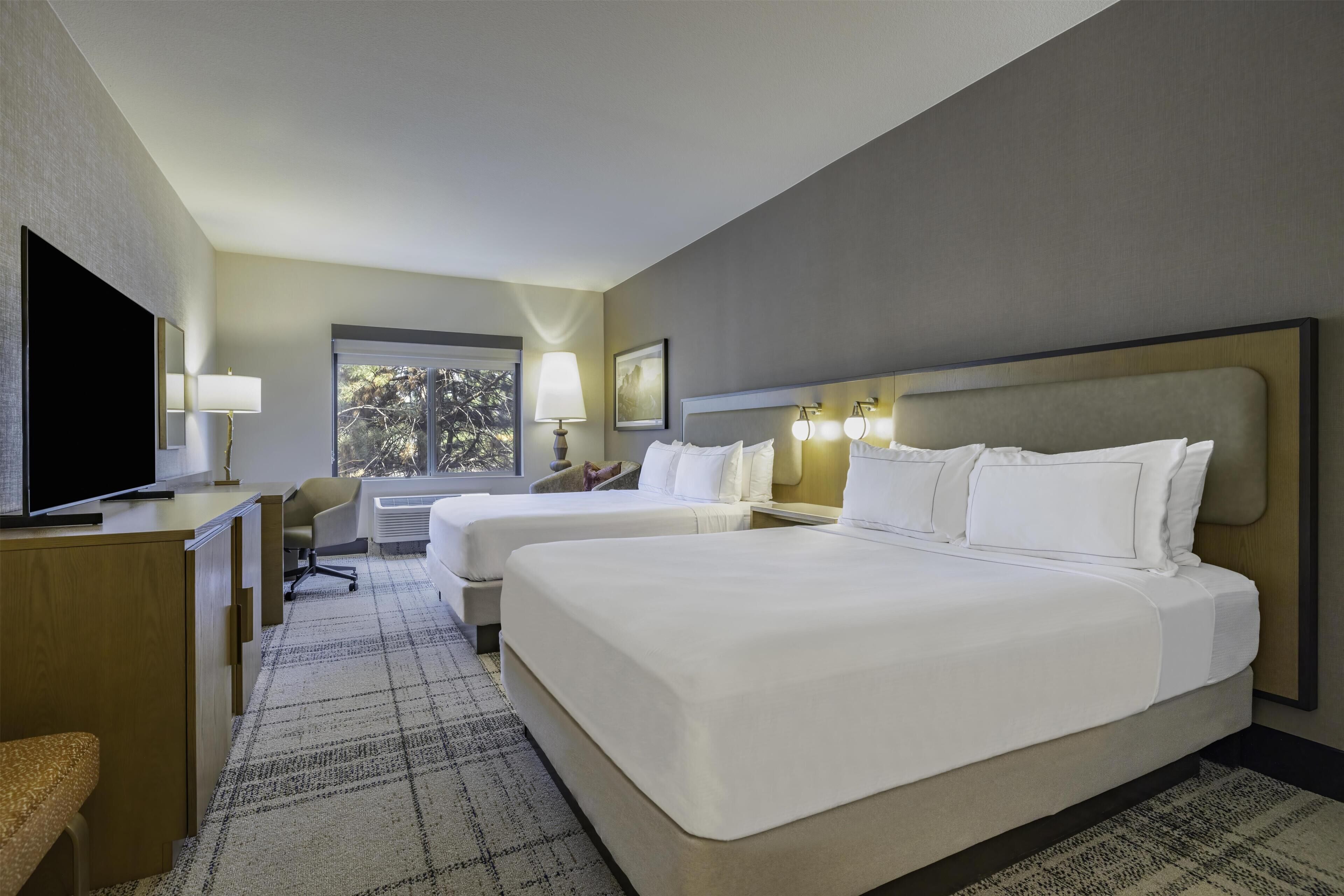 A hotel room with two beds, a desk, a chair, a TV, and a window with a view of trees.