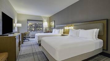 A hotel room with two beds, a desk, a chair, a TV, and a window with a view of trees.