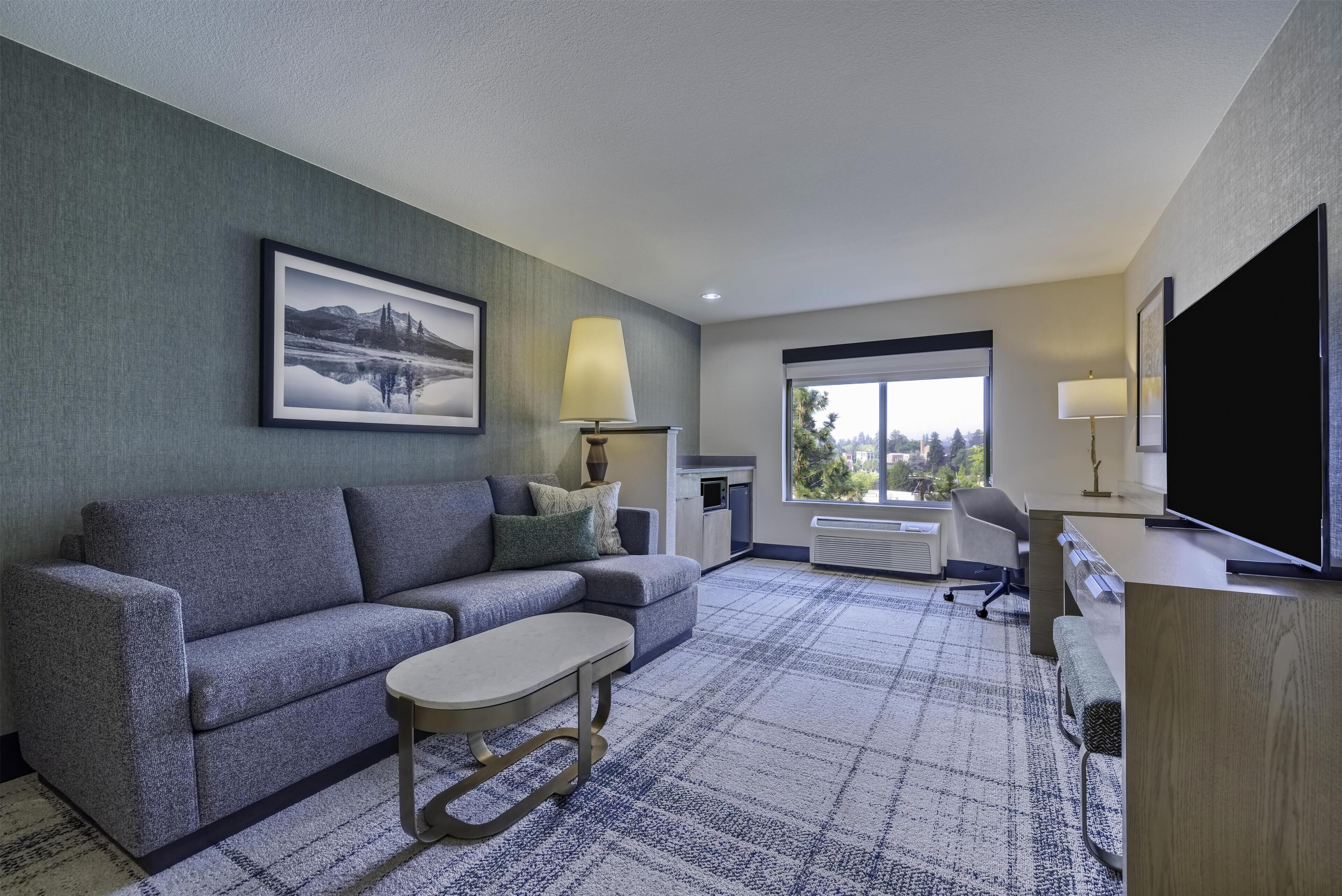 A modern hotel room with a sectional sofa, a small table, a TV, and a large window offering a city view.