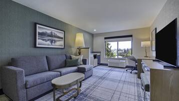 A modern hotel room with a sectional sofa, a small table, a TV, and a large window offering a city view.