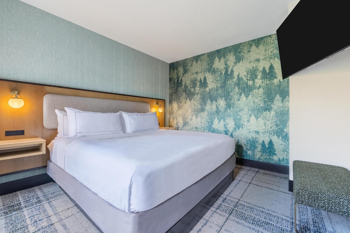 Quarto de hotel com cama, mesa de cabeceira, luminária fixada na parede e papel de parede com tema de floresta.