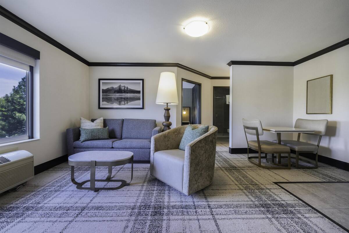 Quarto de hotel com sofá, poltrona, mesa de jantar e cadeiras. Há uma janela com vista, um abajur e um quadro na parede.