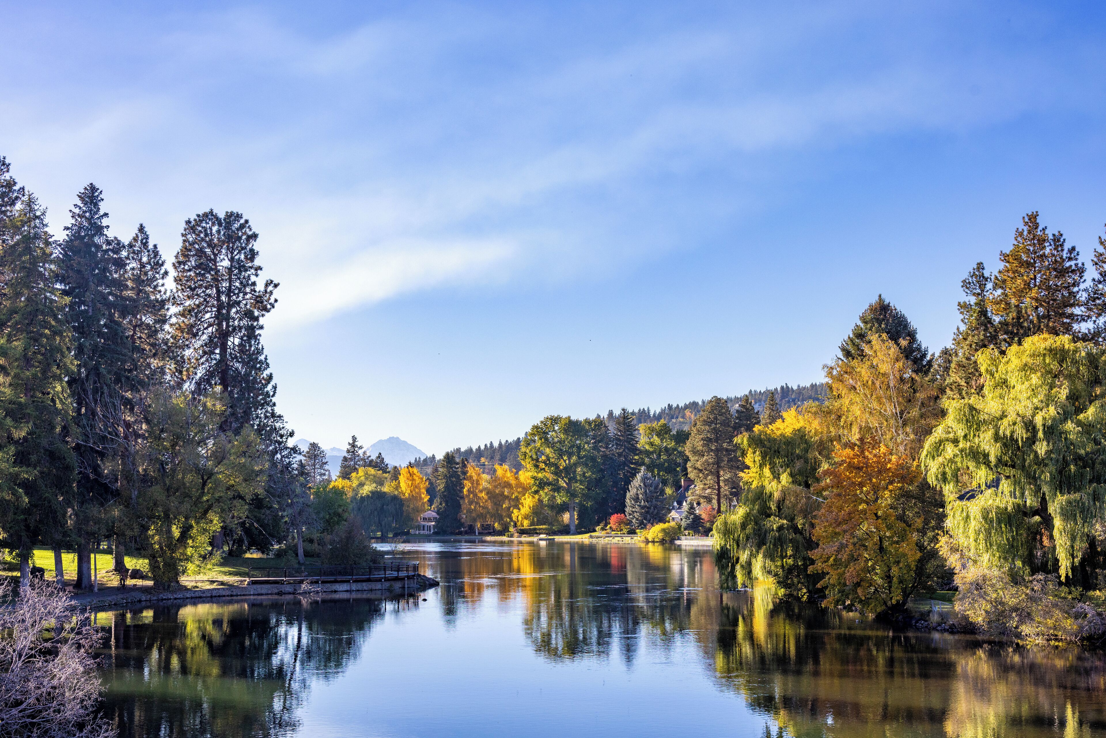 A serene lakeside scene with trees reflecting on the water, a gazebo, and a clear sky.