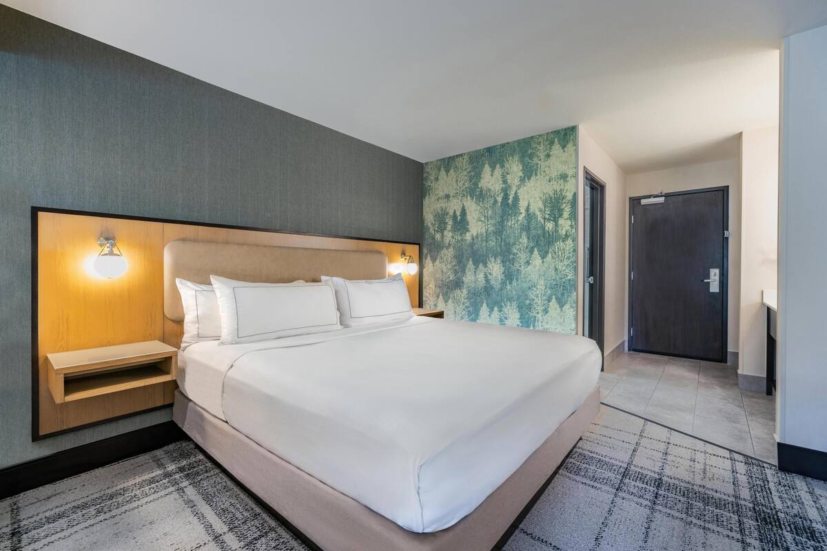 Quarto de hotel com uma cama grande, mesas de cabeceira, cabeceira com iluminação embutida, parede estampada e uma porta.
