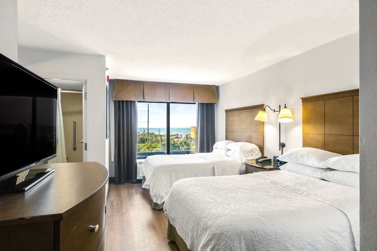 A hotel room with two beds, a TV, and a view of the ocean.