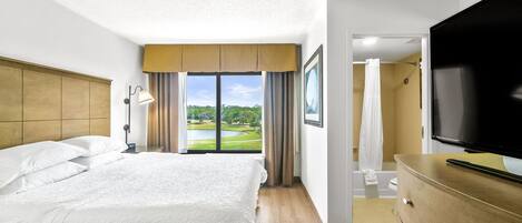 A hotel room with a large bed, a flat-screen TV, a wooden dresser, and a view of a golf course through the window.