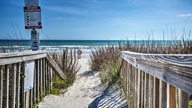 A wooden boardwalk leading to a beach with a flag warning system and a lifebuoy.