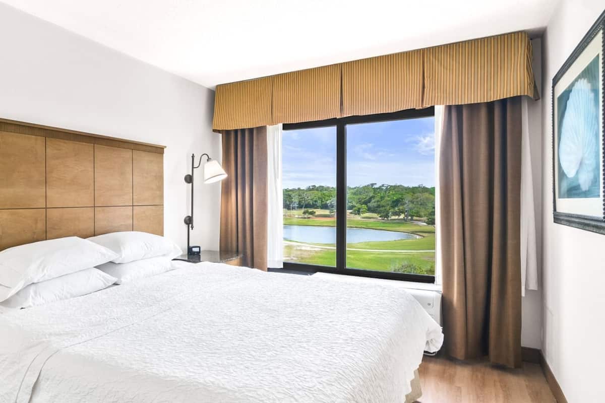 A hotel room with a large bed, a nightstand, a lamp, a window with a view of a golf course, and a framed picture on the wall.