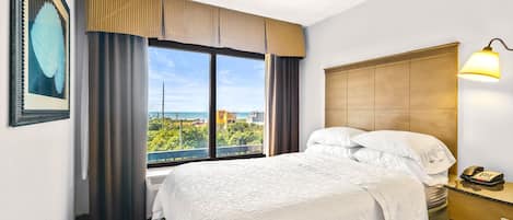 A hotel room with a large window, a bed with white linens, a bedside table with a lamp, and a framed picture on the wall.