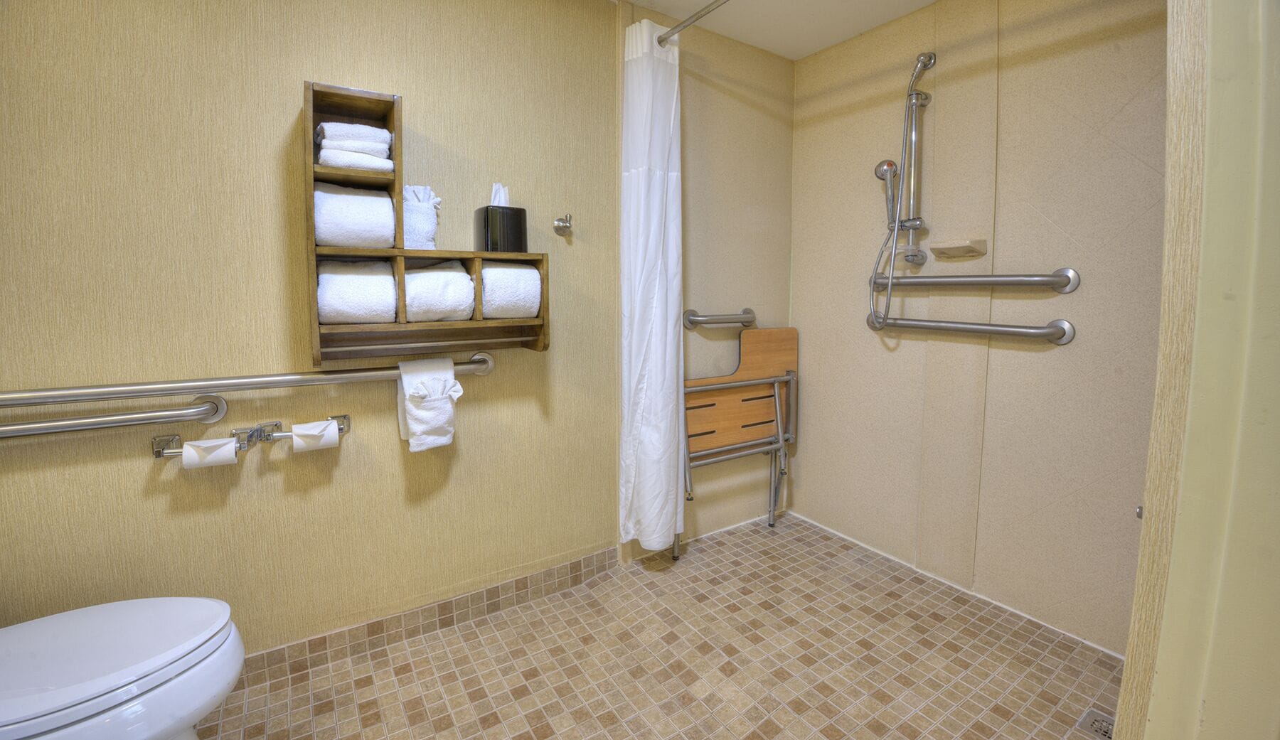A bathroom with a toilet, a shelf with towels, a shower with grab bars, and a shower curtain.