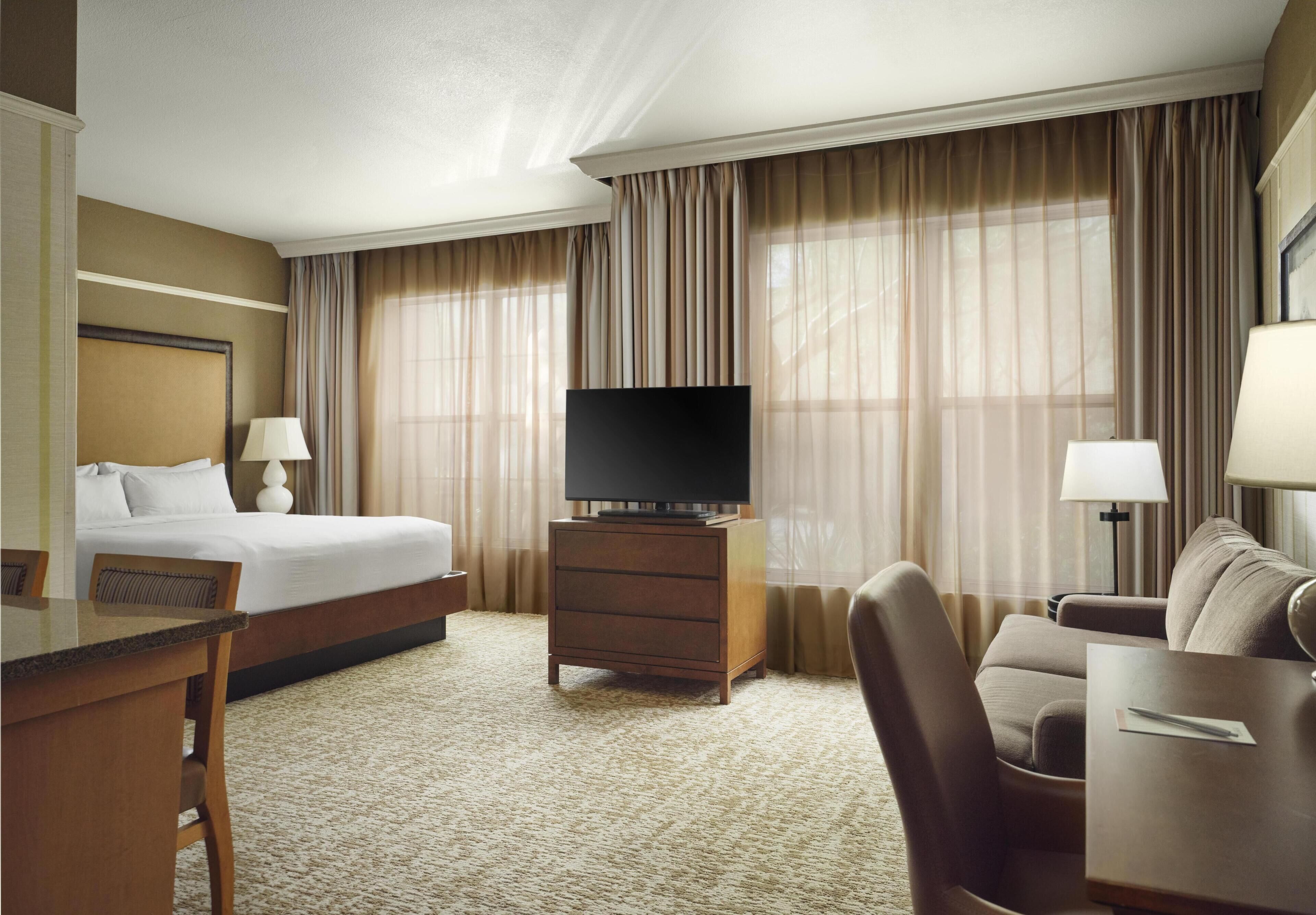 A hotel room with a bed, a TV on a wooden stand, a desk with a chair, a sofa, and a lamp.