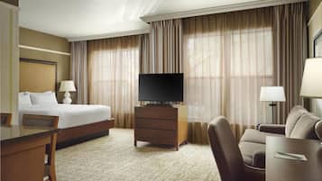 Habitación de hotel con cama, televisor sobre un mueble de madera, escritorio con silla, sofá y lámpara.