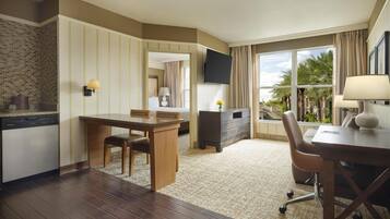 Habitación de hotel con una mesa de comedor de madera, un sofá, un televisor montado en la pared y vistas a palmeras a través de la ventana.