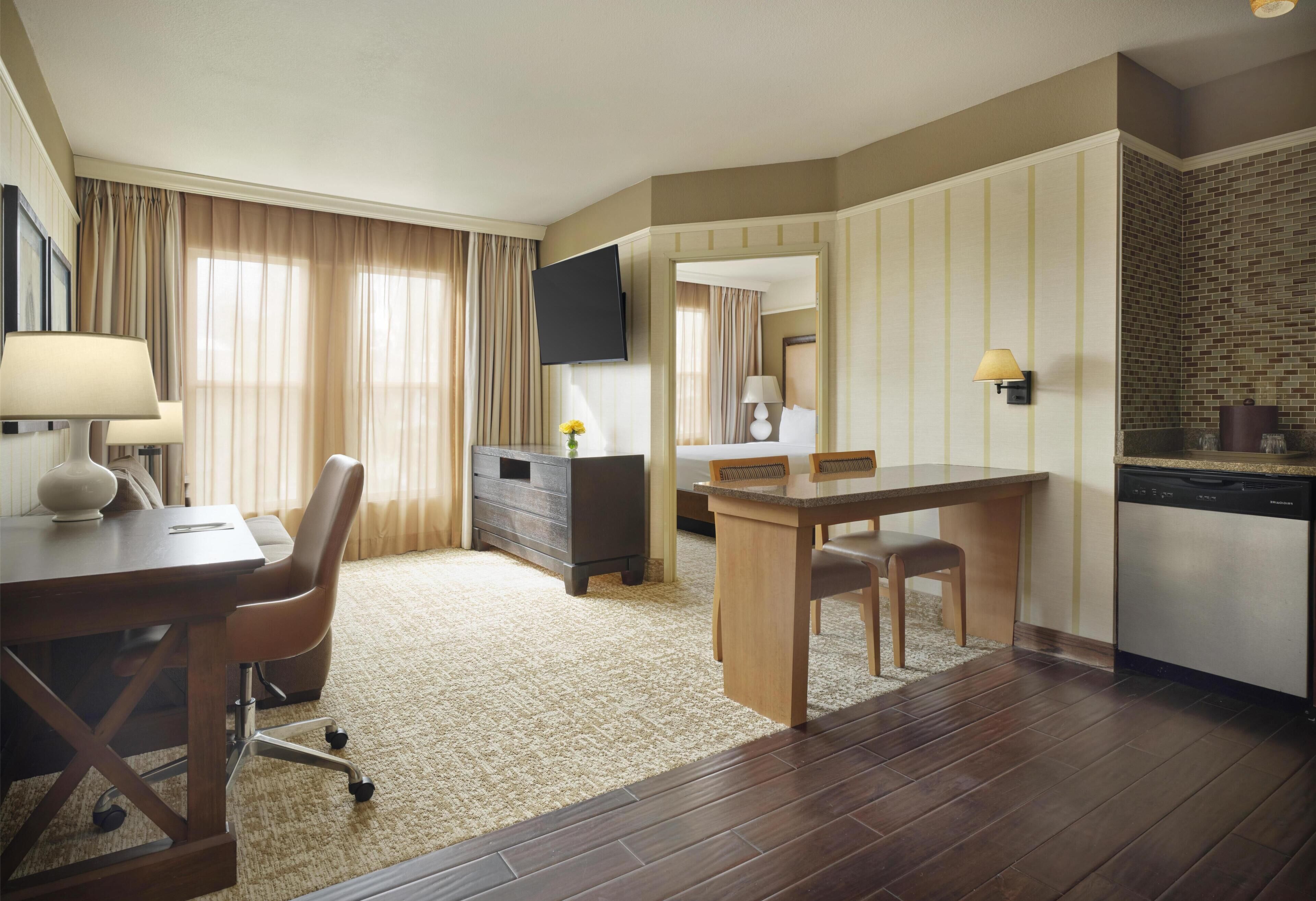A hotel room with a desk, a chair, a dining table, a TV, a dresser, a mirror, a lamp, a rug, and a dishwasher.