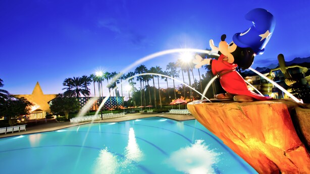 Scena notturna in un parco acquatico con una grande piscina, palme e una statua di Topolino.