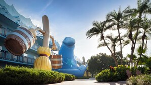 Un'ampia statua blu raffigura un personaggio che tiene in mano una scopa e un secchio, con un edificio sullo sfondo.