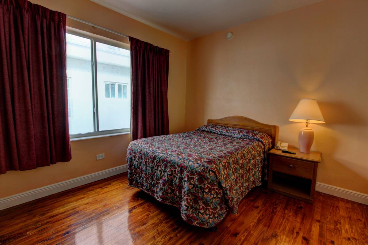 Une chambre avec un plancher en bois, un lit recouvert d’une couvre-lit à motifs, une table de chevet avec une lampe et une fenêtre avec des rideaux.
