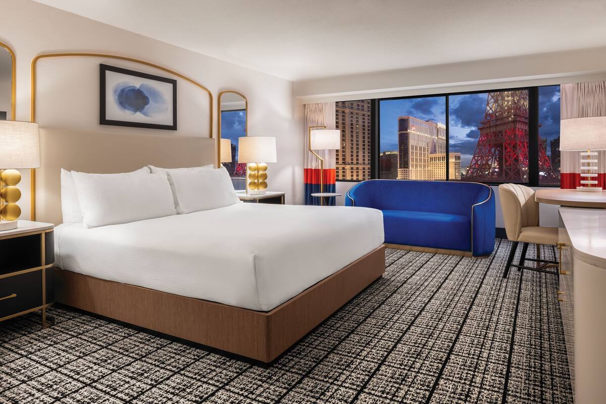 Habitación de hotel con una cama grande, un sofá azul, un escritorio y vistas a la Torre Eiffel.