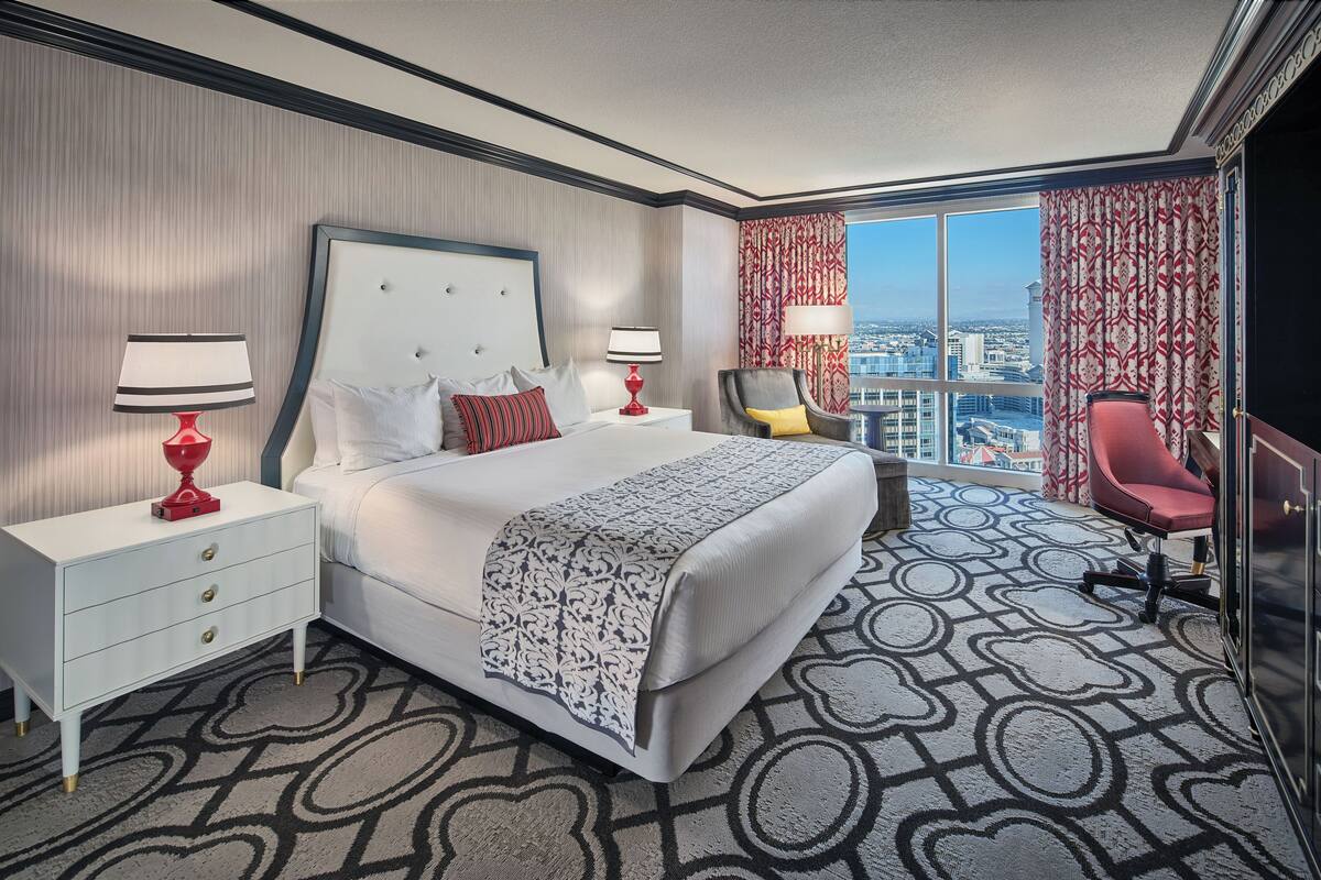 Habitación de hotel con una cama grande, dos mesitas de noche con lámparas, una silla roja y vistas de la ciudad a través de amplias ventanas.