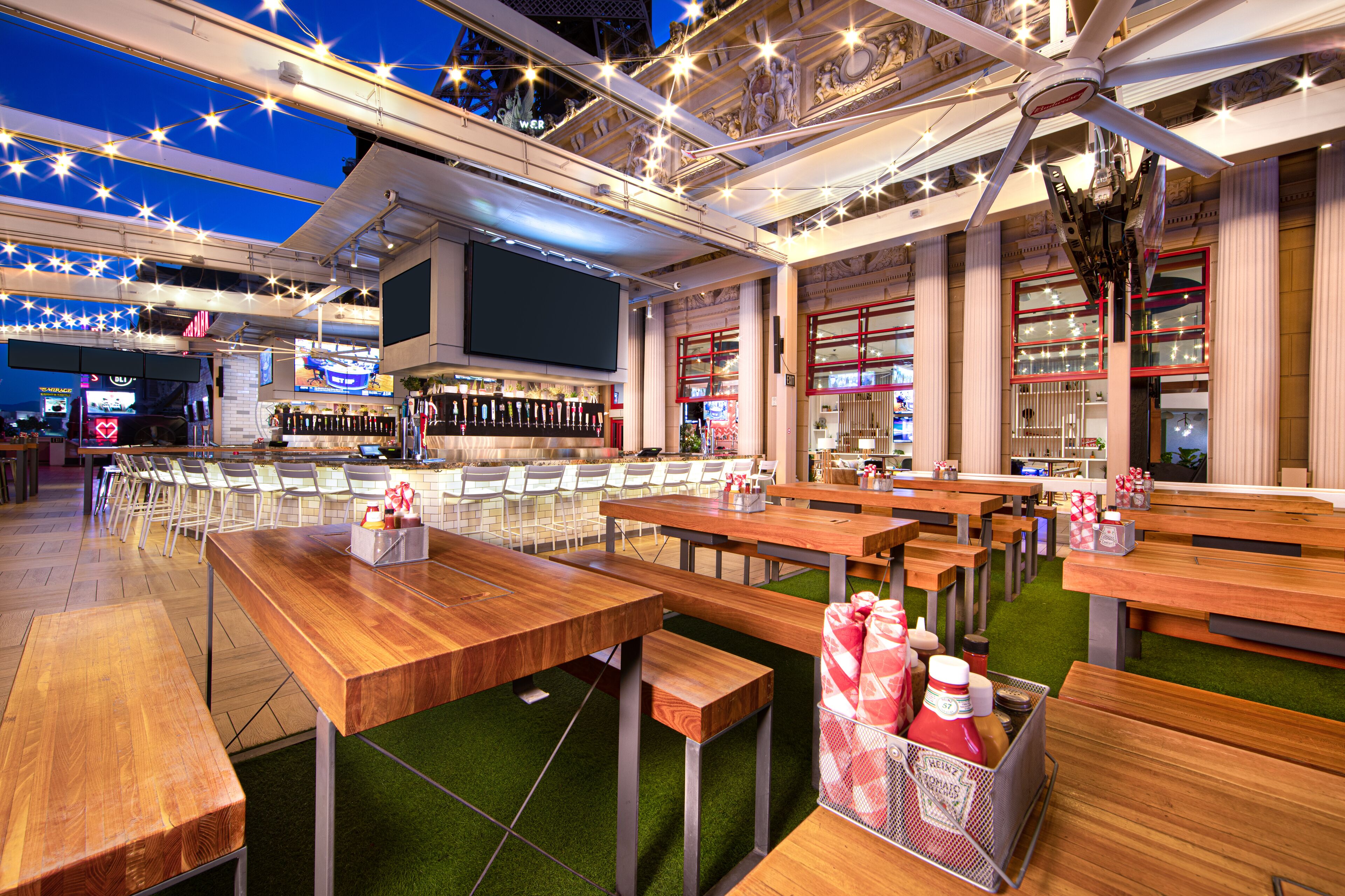 Un restaurante con mesas y bancos de madera, una zona de bar y una pantalla grande.