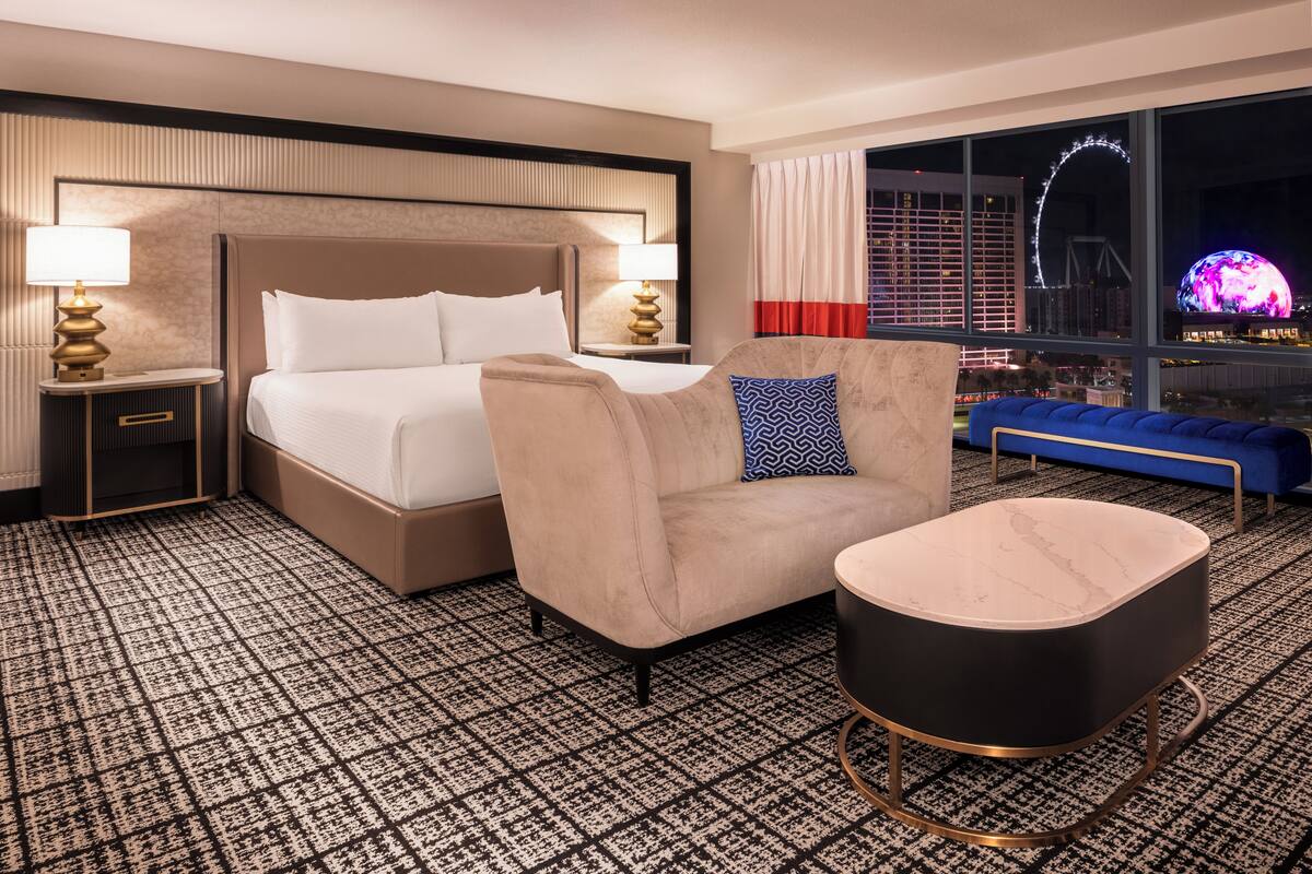 Habitación de hotel con una cama grande, un sofá, una mesita y vistas a un paisaje urbano con noria.