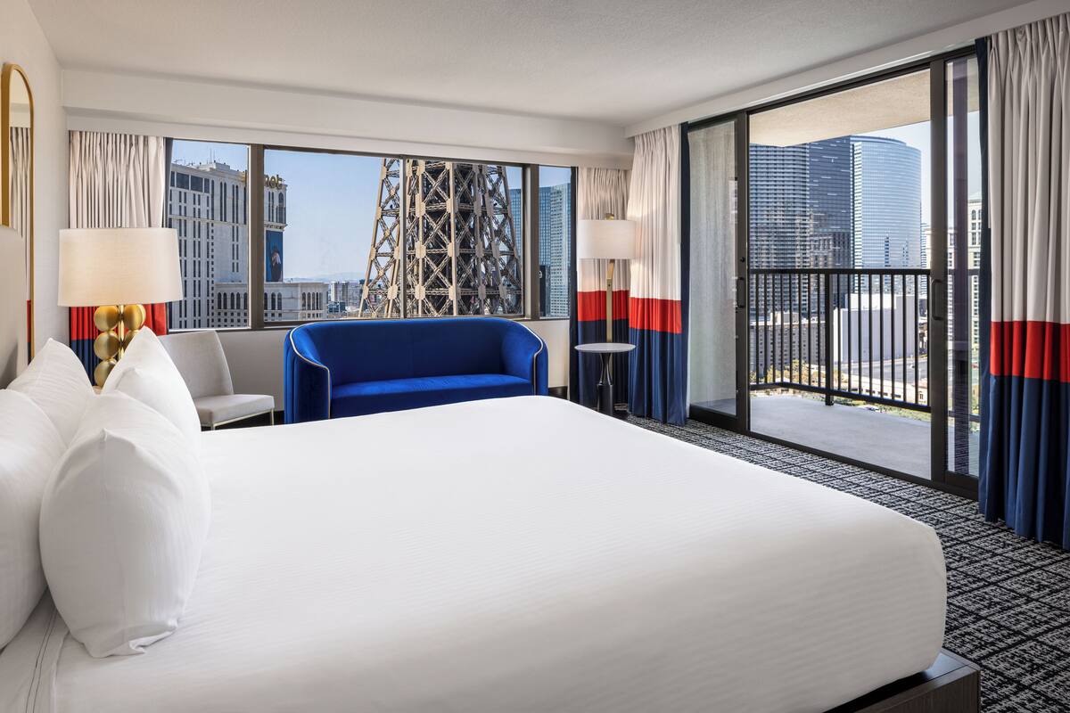 Habitación de hotel con una cama grande, un sillón azul, una mesita con una lámpara y vistas a la ciudad a través de amplios ventanales.