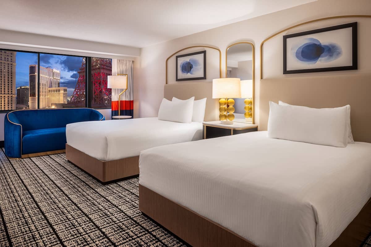 Habitación de hotel con dos camas, un sillón azul, un ventanal grande con vista a la ciudad y una alfombra estampada.