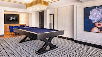 Une table de billard dans une pièce dotée d’un tapis à motifs, d’un lustre, d’un tableau encadré au mur et d’une chaise bleue.