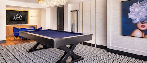 A pool table in a room with a patterned carpet, a chandelier, a framed picture on the wall, and a blue chair.