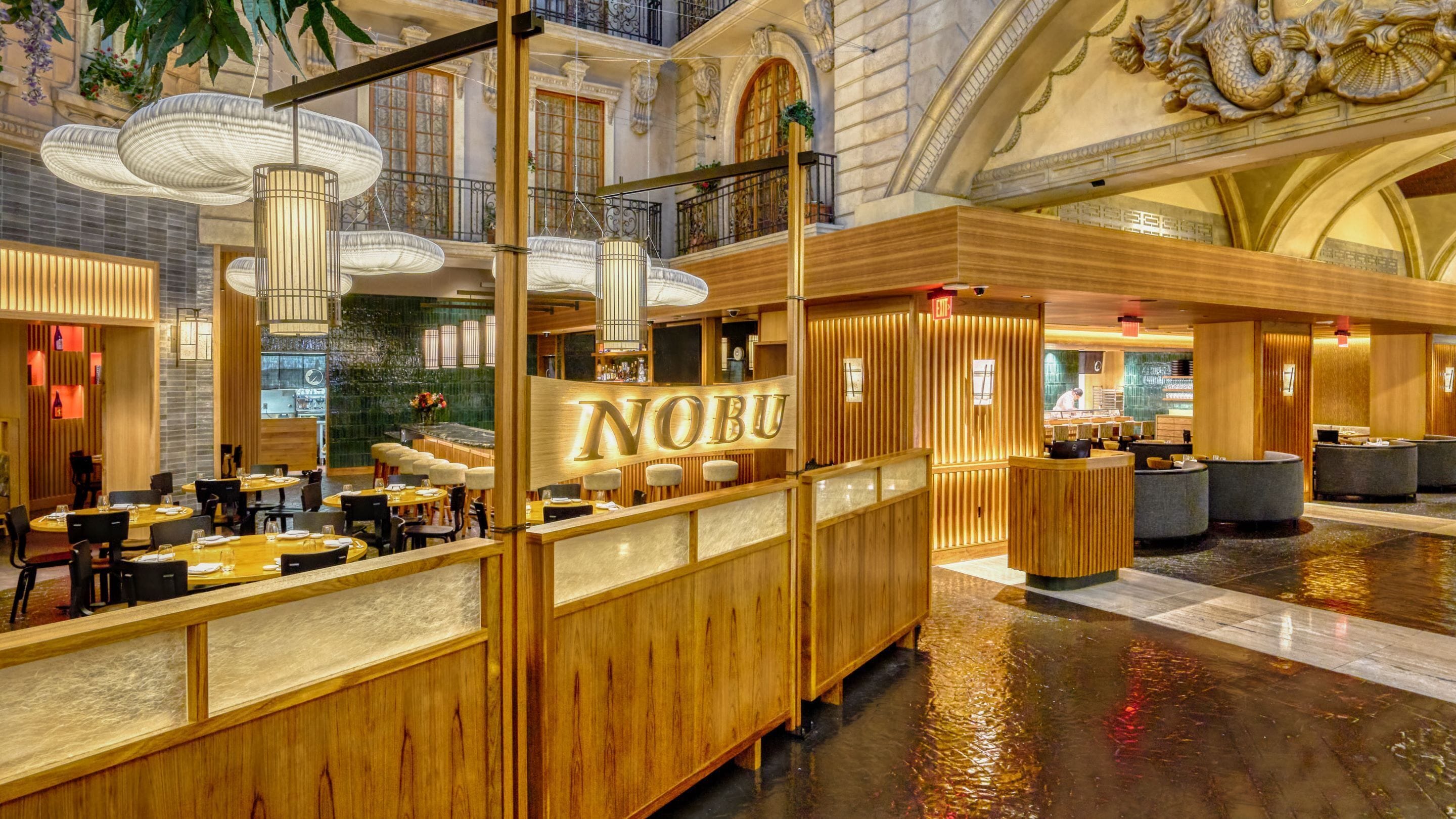 A spacious lobby with a wooden reception desk labeled "NOBU," multiple seating areas, and a high ceiling with decorative elements.