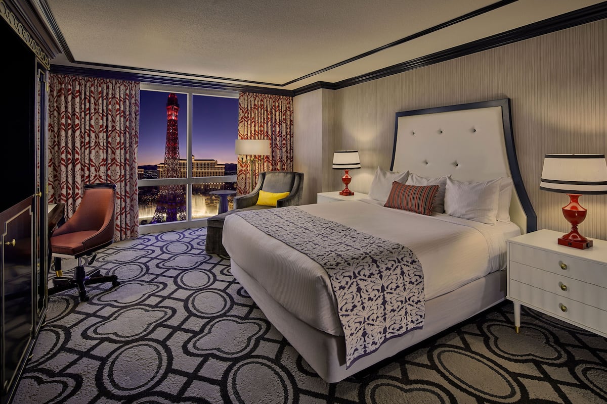 Habitación de hotel con una cama grande, dos mesitas de noche, una silla, un escritorio y vistas a un paisaje urbano con una torre.