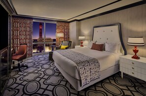 A hotel room with a large bed, two bedside tables, a chair, a desk, and a view of a cityscape with a tower.