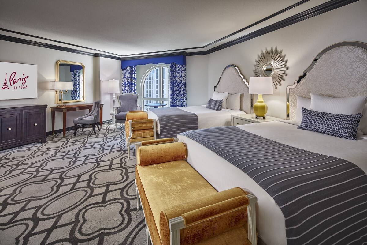 Habitación de hotel con dos camas, un escritorio, una silla y un ventanal con cortinas azules.
