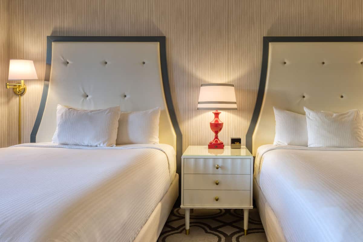 Habitación de hotel con dos camas, cada una con cabecero blanco y almohadas a juego. Hay una mesita de noche entre las camas con una lámpara roja y un reloj digital.