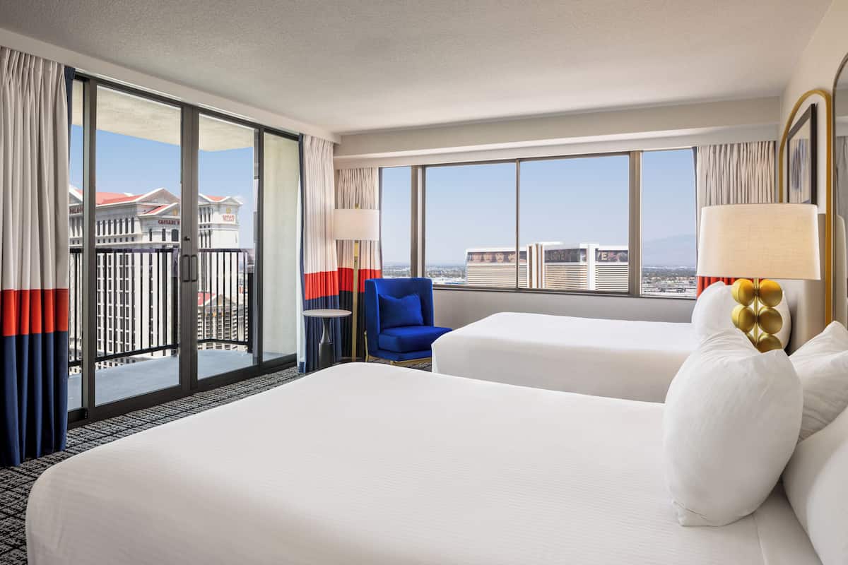 Habitación de hotel con dos camas, una silla azul y grandes ventanales con vistas a la ciudad.