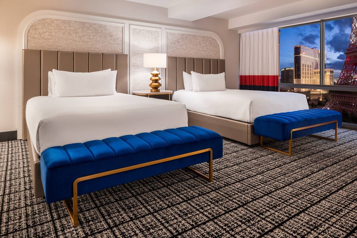 Habitación de hotel con dos camas, un banco azul, alfombra estampada y vista a un paisaje urbano con una torre.