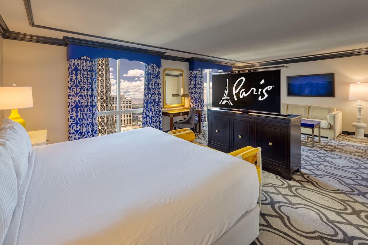 Una habitación de hotel con una cama grande, un escritorio, un televisor mostrando 'París' y vistas a la ciudad.